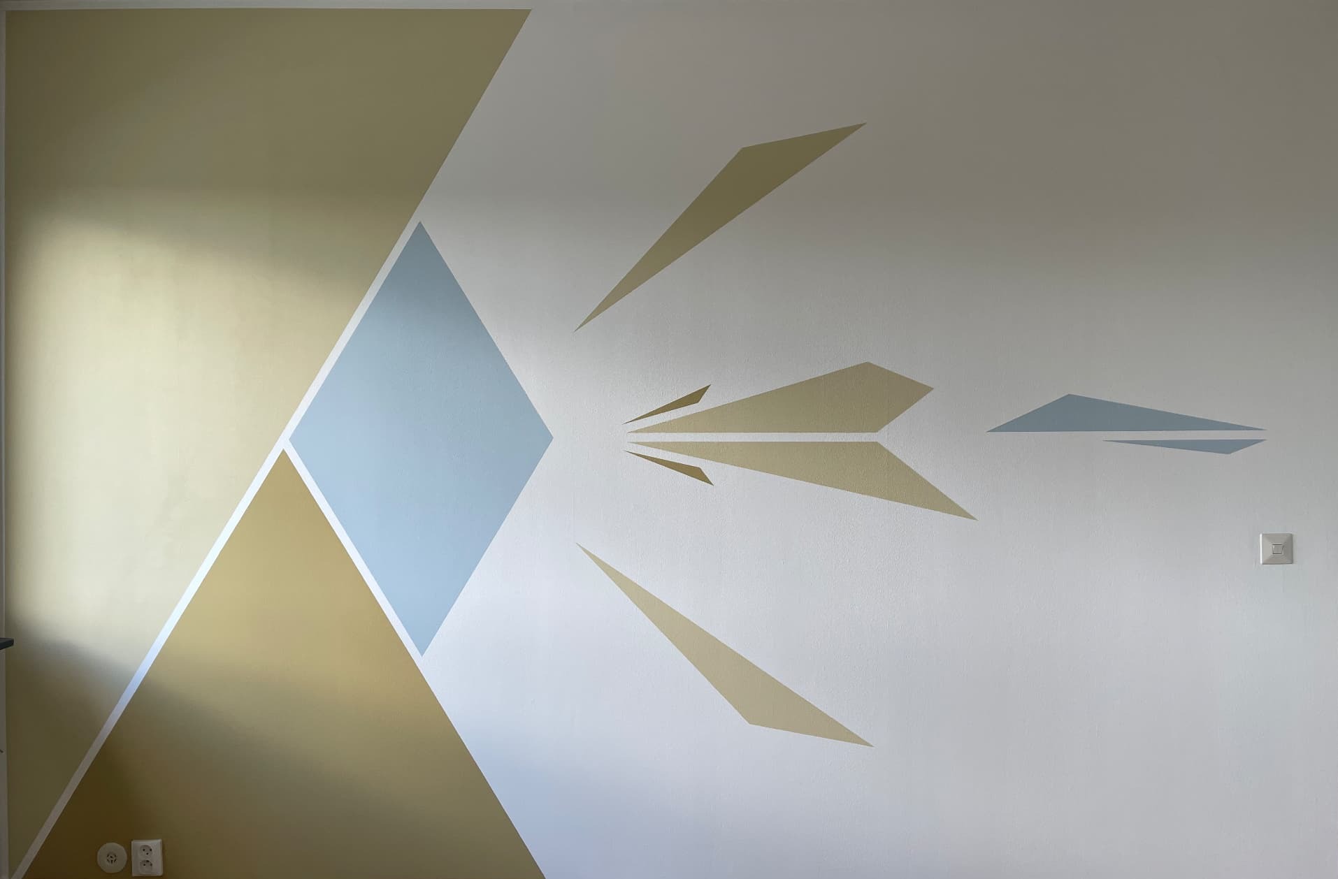 Den första — a geometric wall mural by Driton Memedoski, muted gold and pale blue shapes on an off-white plaster wall